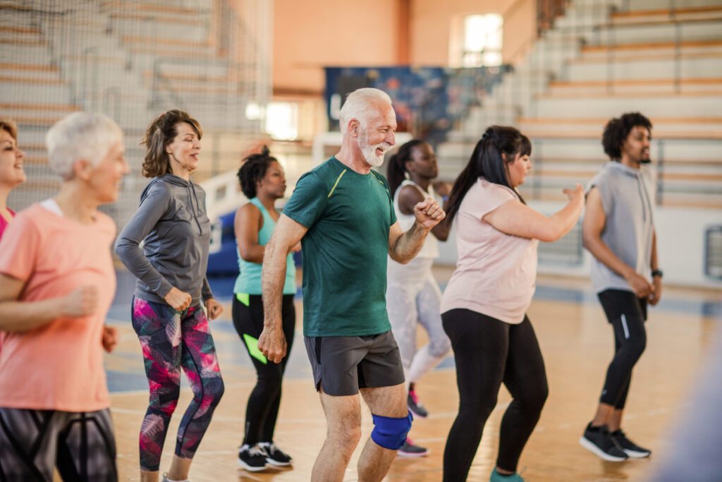 A diverse group of adults of various ages and body types participate in a lively group exercise class in a gymnasium, smiling and moving energetically together.