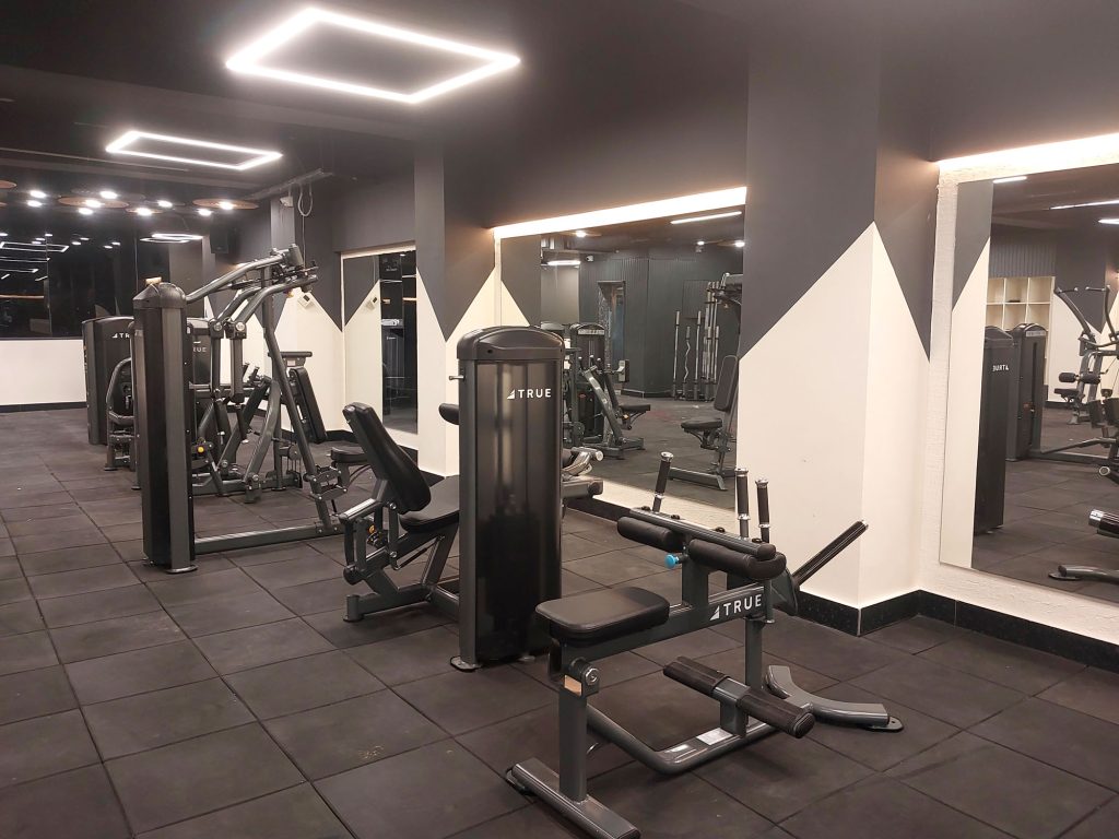 Modern gym with black floors and walls, various exercise machines, mirrors on the walls, and bright square ceiling lights. The space appears clean, spacious, and well-lit, with no people present.