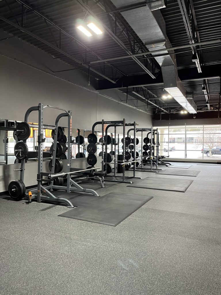 A spacious gym with several squat racks, weight plates, and barbells lined up on rubber mats. Large windows let in natural light, and the area appears clean and organized, with a view of cars outside.