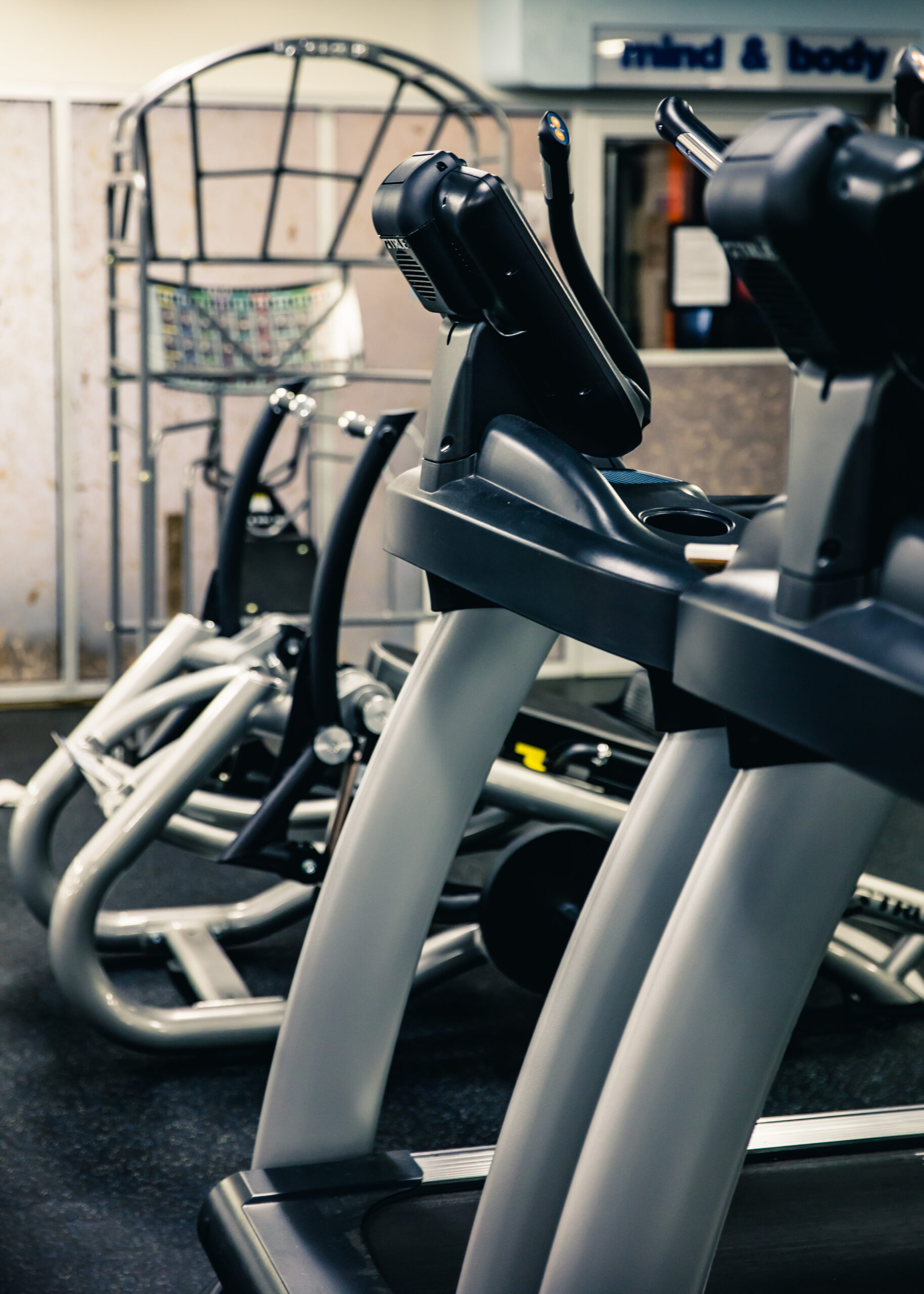 Several treadmills and exercise machines are lined up in a gym with dark flooring and beige walls. The area appears clean and organized, with fitness equipment ready for use.
