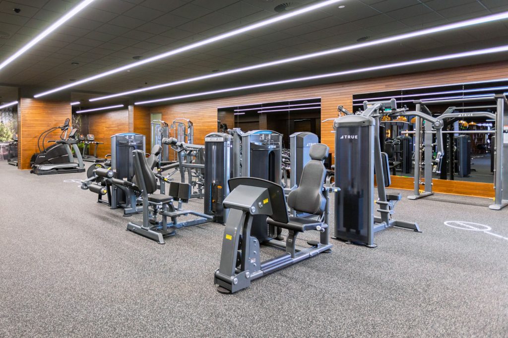 A modern gym with various workout machines, including strength training and cardio equipment, on a carpeted floor with large mirrors and wood-paneled walls. The space is brightly lit with overhead lights.