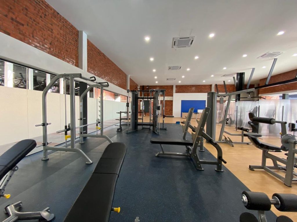 A spacious gym with various exercise machines, weight benches, and fitness equipment on blue and wooden flooring, surrounded by large mirrors and brick walls under bright ceiling lights.