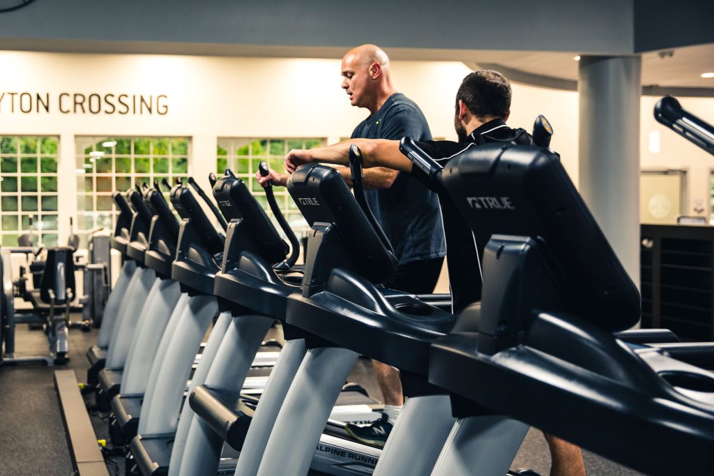 Two men exercise on treadmills in a well-lit gym. One is bald and wearing a blue shirt, the other has dark hair and wears a black shirt. Several treadmills are lined up in the foreground.