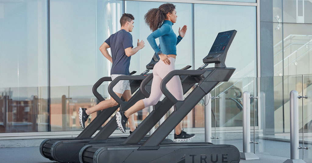 A man and a woman are running on treadmills in a modern gym with large glass windows, letting in natural light and offering a city view in the background.