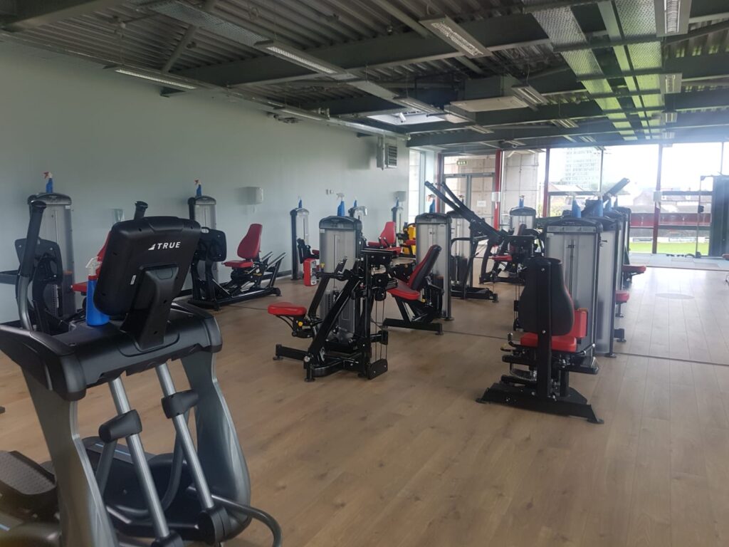 A modern gym with various exercise machines, including ellipticals and weight machines, arranged on a wooden floor. Large windows let in natural light, and the ceiling is industrial-style with exposed pipes.